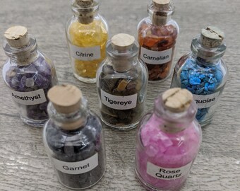 May include: Eight small glass bottles filled with various coloured crystals. Each bottle has a cork stopper and a label identifying the type of crystal: Amethyst, Citrine, Tigerseye, Carnelian, Turquoise, Garnet, Rose Quartz.