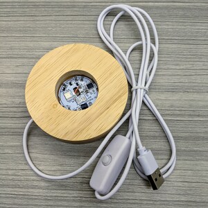 May include: A wooden base with a circular cutout for displaying a small object. The base has a white USB cable attached with an inline switch. The base is designed to be used with a USB power source.
