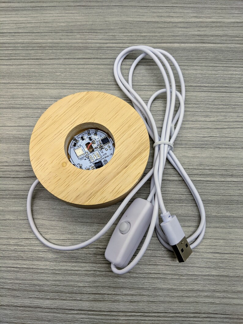 May include: A wooden base with a circular cutout for displaying a small object. The base has a white USB cable attached with an inline switch. The base is designed to be used with a USB power source.