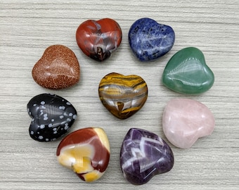 May include: A collection of ten heart-shaped polished stones, each a different colour and type of crystal. The stones are arranged in a circle on a light wood surface.