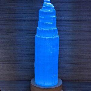 May include: A blue selenite crystal lamp with a wooden base. The crystal is shaped like a tower and is illuminated from within, creating a soft blue glow.