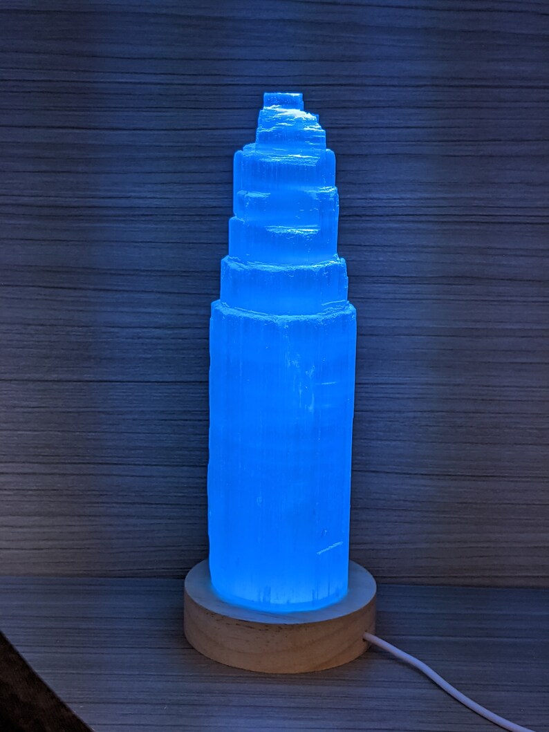May include: A blue selenite crystal lamp with a wooden base. The crystal is shaped like a tower and is illuminated from within, creating a soft blue glow.