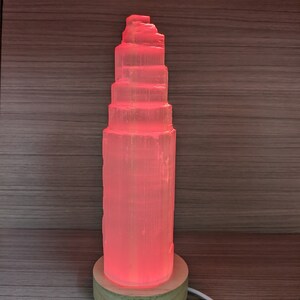 May include: A pink Himalayan salt lamp with a wooden base. The lamp is lit and glowing a warm pink colour.