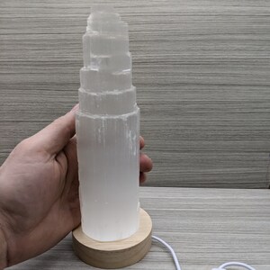 May include: A white selenite crystal lamp with a wooden base and a white cord with an on/off switch. The crystal is stacked in a tower shape.