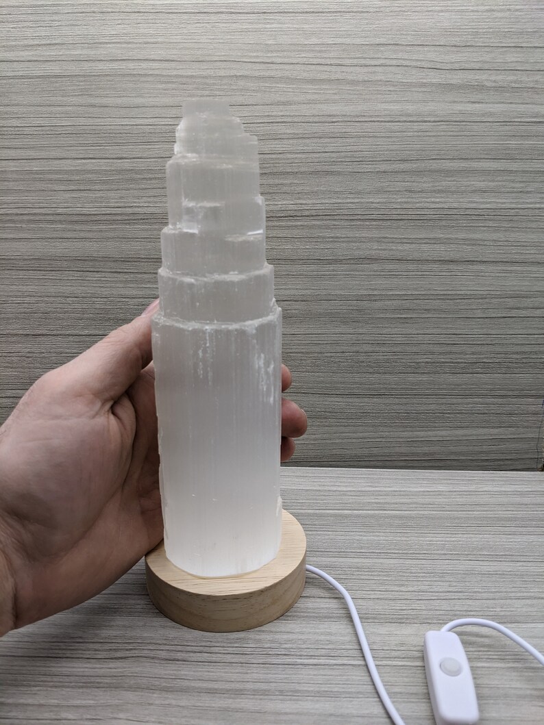 May include: A white selenite crystal lamp with a wooden base and a white cord with an on/off switch. The crystal is stacked in a tower shape.