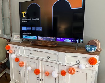 May include: A white dresser with a TV on top. The TV is displaying a screen with the words "Tennessee Volunteers" and "News & Highlights". There is a garland of orange and white pom-poms hanging on the front of the dresser.