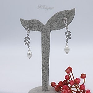 May include: A pair of silver earrings with a pearl drop and a leaf design. The earrings are displayed on a gray stand.