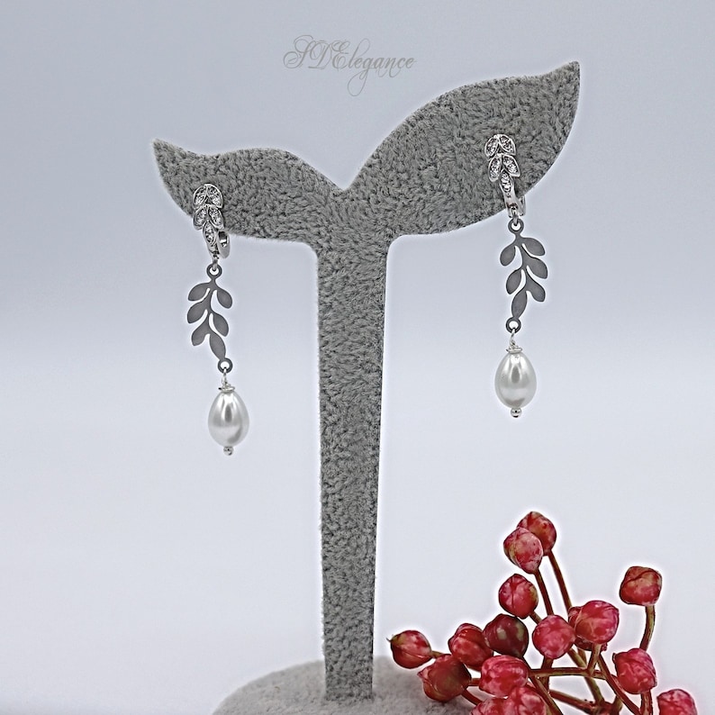 May include: A pair of silver earrings with a pearl drop and a leaf design. The earrings are displayed on a gray stand.