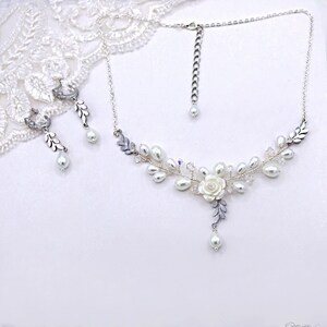 May include: A delicate silver necklace and earrings set with a floral design featuring white pearls and crystal accents. The necklace has a pendant with a white flower and leaves.