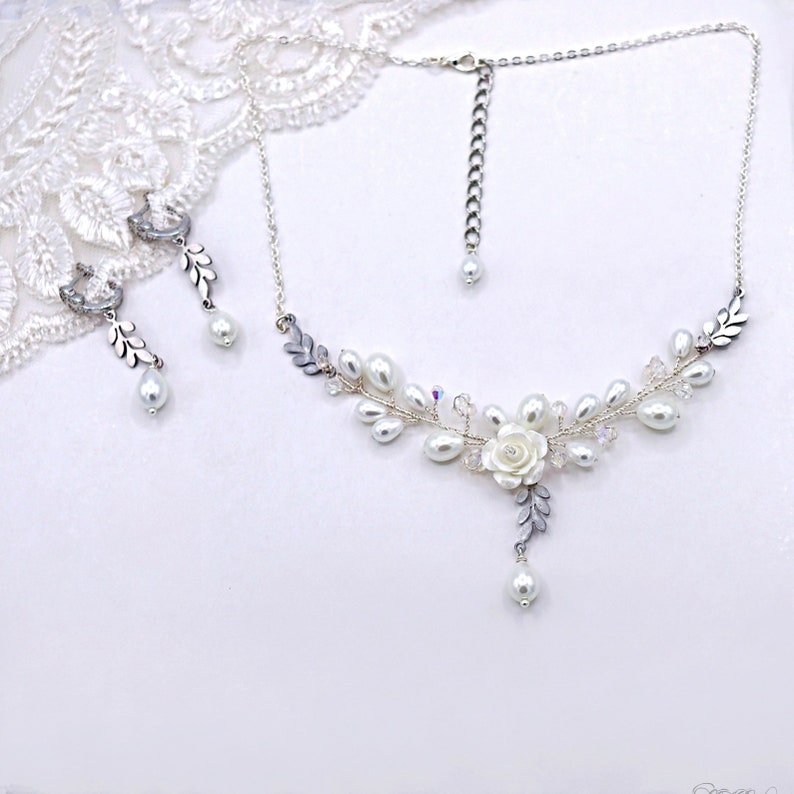 May include: A delicate silver necklace and earrings set with a floral design featuring white pearls and crystal accents. The necklace has a pendant with a white flower and leaves.