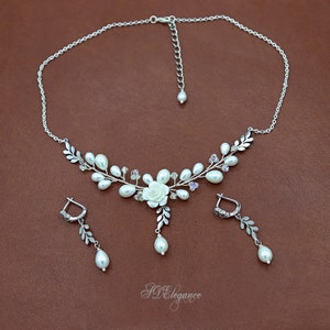 May include: A delicate silver necklace and earrings set with a floral design featuring white pearls, crystal accents, and a white flower charm. The necklace has a delicate chain and a floral pendant with a white pearl drop.
