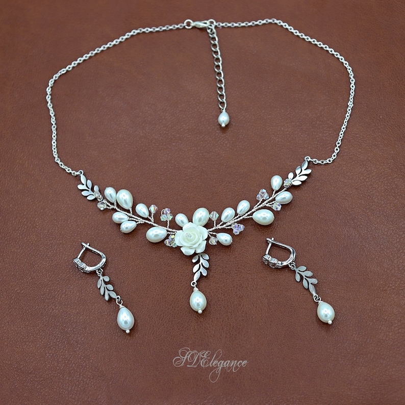 May include: A delicate silver necklace and earrings set with a floral design featuring white pearls, crystal accents, and a white flower charm. The necklace has a delicate chain and a floral pendant with a white pearl drop.