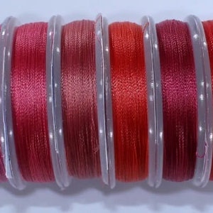May include: Ten spools of thread in various shades of red, pink, and brown. The spools are arranged in a row from lightest to darkest.
