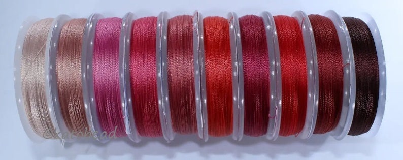 May include: Ten spools of thread in various shades of red, pink, and brown. The spools are arranged in a row from lightest to darkest.