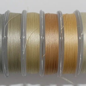 May include: A collection of 10 spools of thread in various shades of beige, cream, and gray. The thread is wound around clear plastic spools with labels.
