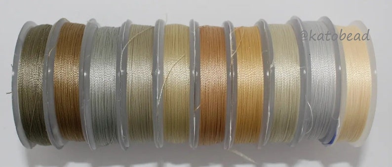May include: A collection of 10 spools of thread in various shades of beige, cream, and gray. The thread is wound around clear plastic spools with labels.