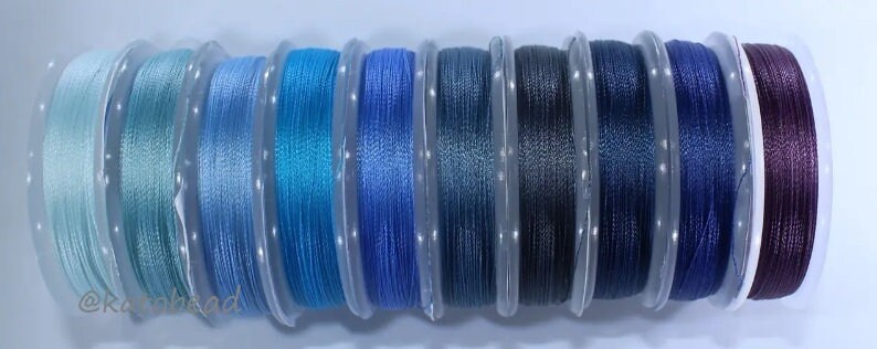 May include: Assortment of 10 spools of thread in various shades of blue and purple. The thread is wound around clear plastic spools.