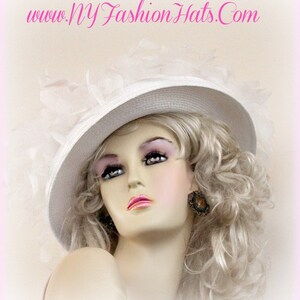 May include: A white hat with a wide brim and a white feather trim. The hat is worn by a mannequin with blonde hair and a light pink blush.