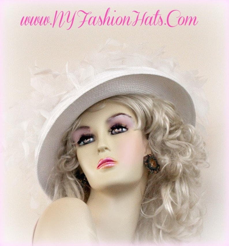 May include: A white hat with a wide brim and a white feather trim. The hat is worn by a mannequin with blonde hair and a light pink blush.
