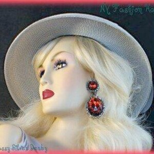 May include: A silver derby hat with a wide brim and a simple design. The hat is being worn by a mannequin with blonde hair and red earrings. The text "Classy Silver's Derby" is visible at the bottom of the image.