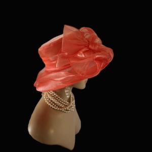 May include: A coral-coloured hat with a large bow and a wide brim. The hat is made of a sheer, flowing fabric and is displayed on a mannequin head with a pearl necklace. The background is black.