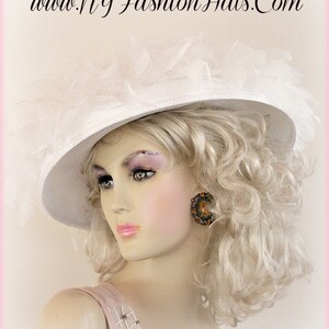 May include: A white wide-brimmed hat with a large white feather trim. The hat is worn by a mannequin with blonde curly hair and a pink dress.