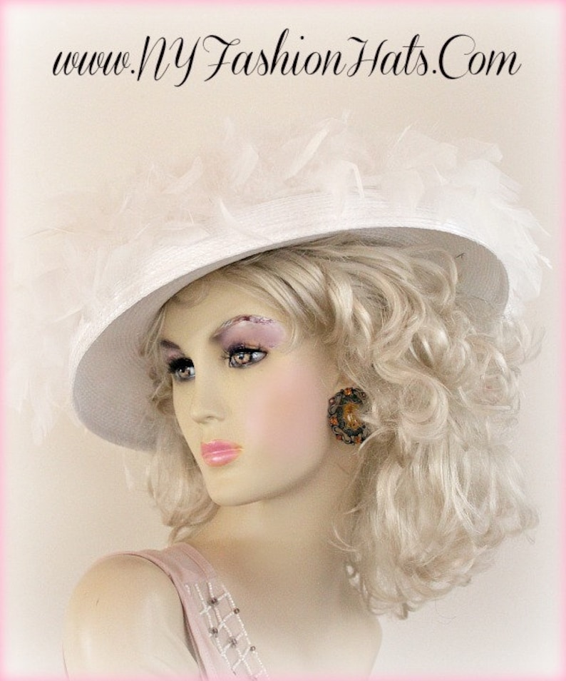 May include: A white wide-brimmed hat with a large white feather trim. The hat is worn by a mannequin with blonde curly hair and a pink dress.