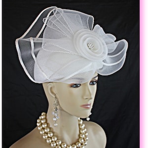 May include: A white, wide-brimmed hat with a large, white flower and a rhinestone center. The hat is made of a delicate, sheer fabric and has a ruffled design.