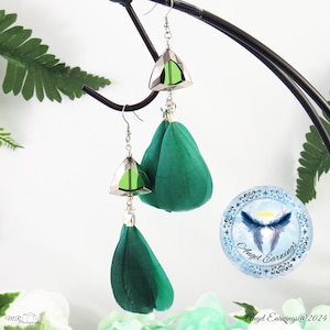 Puede incluir: Un par de pendientes colgantes con detalles de plumas verdes y abalorios de cristal verde triangulares. Los pendientes están colgados de un soporte negro. El texto "Angel Earrings" es visible en un círculo azul.