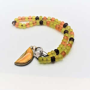 May include: A necklace featuring a double strand of faceted beads in shades of lime green, orange, and dark brown. The necklace includes a semi-circular pendant with a banded yellow and brown pattern, attached to a silver-toned textured clasp.