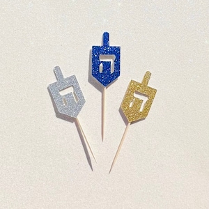 May include: Three glitter-covered dreidels on wooden skewers. The dreidels are blue, silver, and gold.