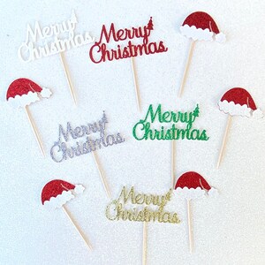 May include: Six cupcake toppers with a festive Christmas theme. Each topper features a red, green, silver, or gold glitter "Merry Christmas" message and a small Santa hat.