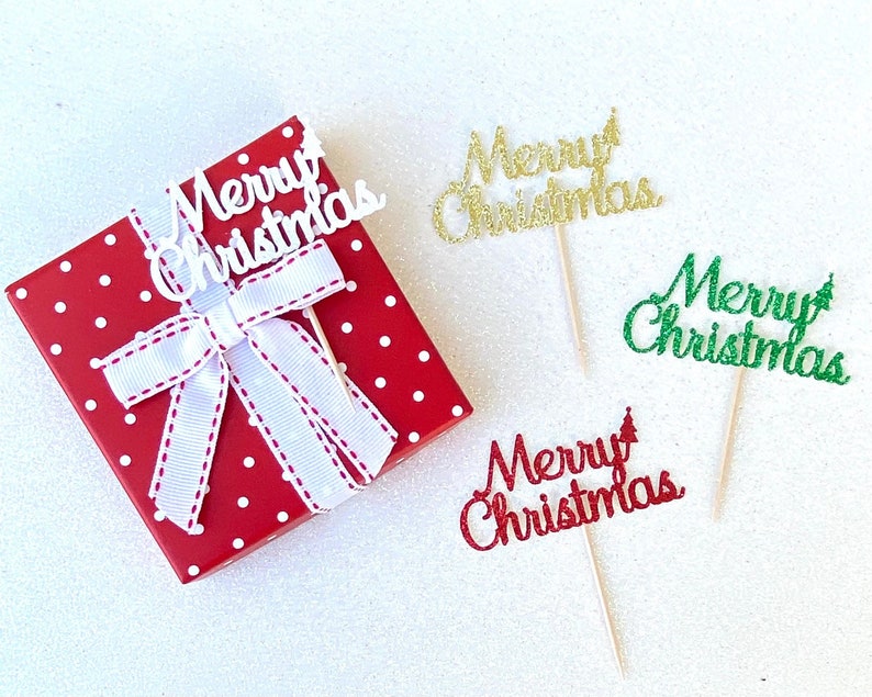 May include: Four "Merry Christmas" cake toppers with different colors and glitter. The toppers are on a white background. One topper is on a red gift box with white polka dots and a white ribbon.