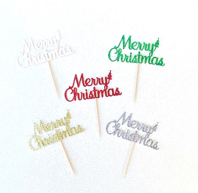 May include: Five cupcake toppers with the words "Merry Christmas" in different colors. The toppers are made of glitter and are attached to wooden sticks.