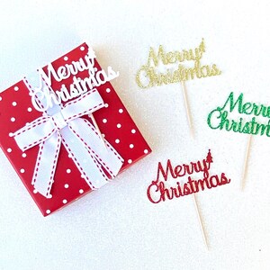 May include: Four "Merry Christmas" cake toppers with a red polka dot gift box. The toppers are made of glitter paper and come in white, gold, green, and red.