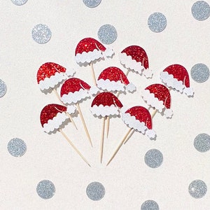 May include: A collection of ten red and white glitter Santa hats on wooden skewers. The hats are arranged in a circle on a white background with silver glitter dots.