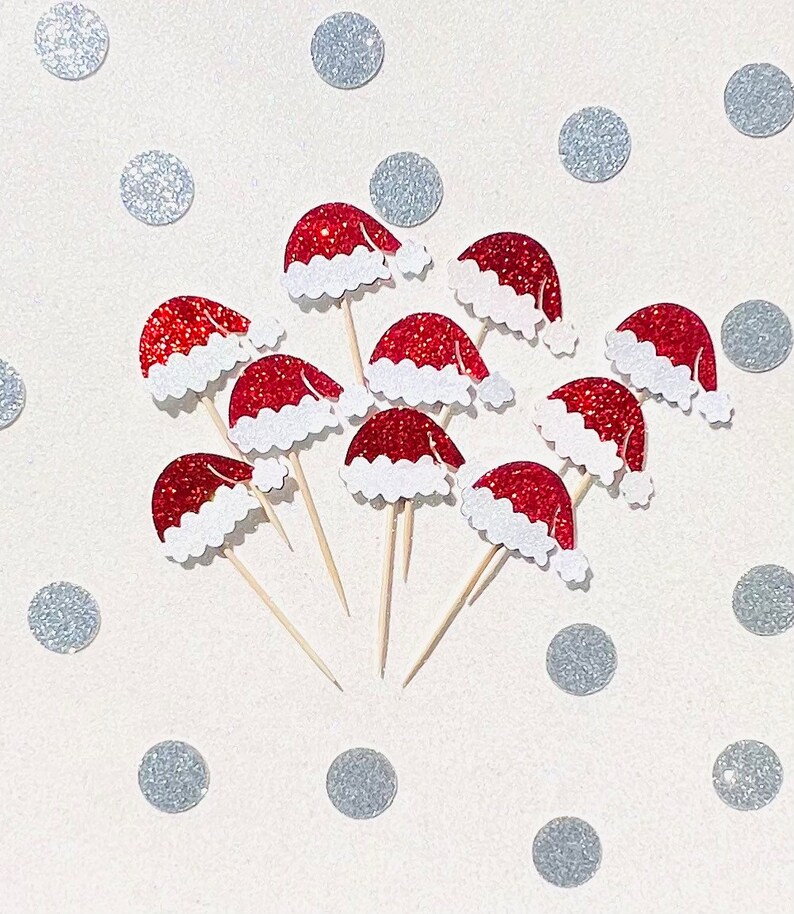 May include: A collection of ten red and white glitter Santa hats on wooden skewers. The hats are arranged in a circle on a white background with silver glitter dots.