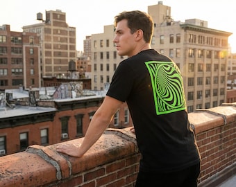Può includere: T-shirt nero con un design grafico verde neon e nero sul retro. Il design è un quadrato con un motivo a spirale a illusione ottica. La persona è in piedi su un muro di mattoni con lo skyline della città sullo sfondo.