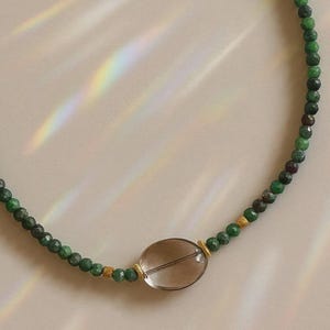 Op de afbeelding: Een ketting met groene en bordeauxrode kralen, met een grote, heldere ovale hanger. De ketting heeft goudkleurige accenten en wordt gepresenteerd tegen een lichte achtergrond.