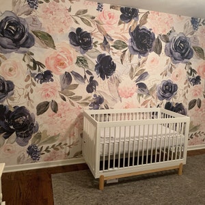 May include: A white wooden crib with a mattress sits against a wall covered in floral wallpaper featuring large navy blue and soft pink roses, green leaves, and delicate stems. The wallpaper pattern covers the entire wall behind the crib.
