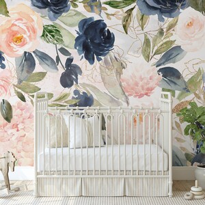May include: A white crib with a white mattress and two pillows sits in front of a wall with a floral mural. The mural features large pink and blue watercolor flowers with gold accents.