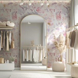 May include: A well-lit boutique with a large arched mirror and clothing racks. The walls are adorned with floral wallpaper in shades of pink, purple, and gray. Various garments hang on the racks, and accessories are displayed on shelves and a white cabinet.