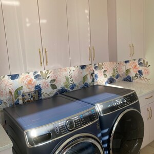 May include: A laundry room featuring a navy blue washer and dryer set. Above, light pink cabinets with gold handles are installed. A floral wallpaper backsplash with pink and blue flowers adds a decorative touch.