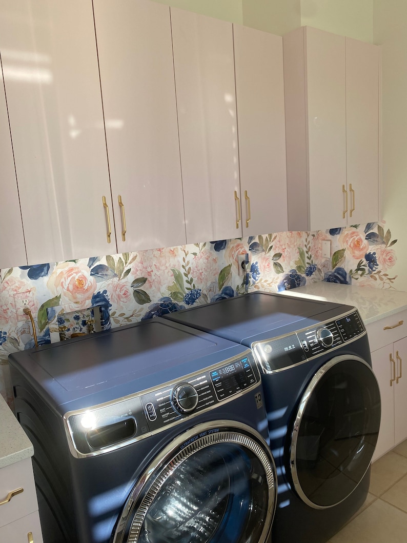 May include: A laundry room featuring a navy blue washer and dryer set. Above, light pink cabinets with gold handles are installed. A floral wallpaper backsplash with pink and blue flowers adds a decorative touch.