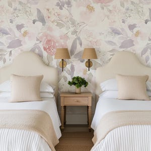 May include: Two twin beds with white bedding, beige headboards, and matching pillows. A small wooden nightstand with a green plant sits between the beds. Floral wallpaper and woven lampshades complete the bedroom decor.