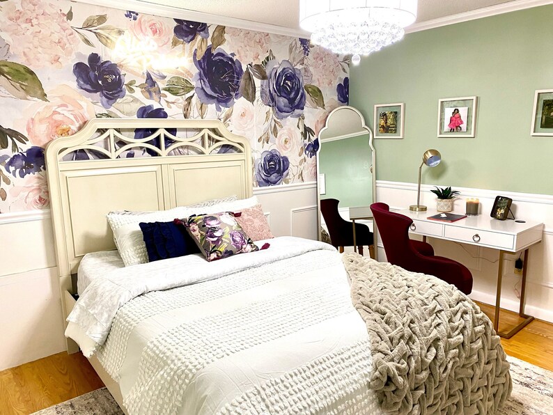 May include: A bedroom with a floral wallpaper and a white bed with decorative pillows. A white desk with a gold lamp and a red chair is in the corner. A large mirror and a chandelier are also visible.