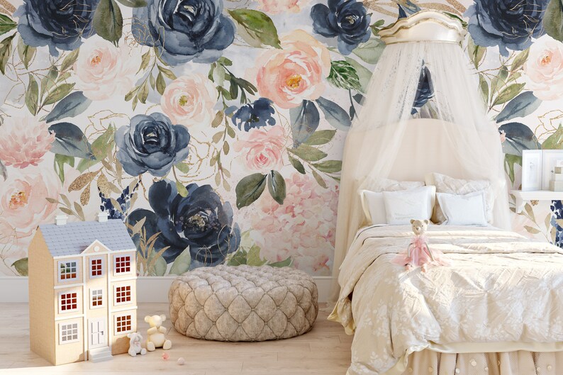 May include: A bedroom scene featuring a floral wallpaper with navy and blush roses. A bed with a canopy, a dollhouse, and a patterned ottoman are in the foreground. A teddy bear sits on the bed. The room has a soft, feminine aesthetic.
