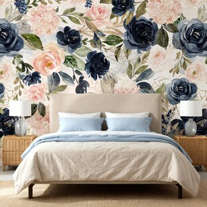 May include: A bedroom scene featuring a beige upholstered bed with light blue bedding, flanked by two wicker nightstands with white lamps. The wall behind the bed is covered in wallpaper with a watercolor floral pattern of navy and blush roses.