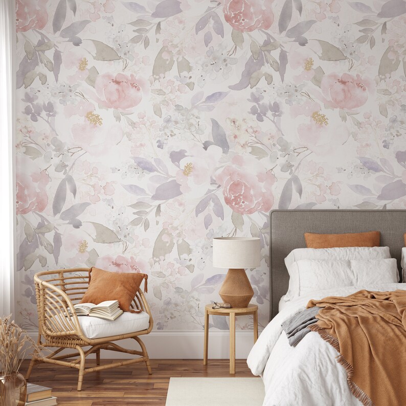 May include: A bedroom scene featuring floral wallpaper in soft pinks, lavenders, and greens. A rattan chair with a burnt orange pillow and a bed with white linens and a brown throw are in the foreground. A wooden side table with a lamp sits beside the bed.