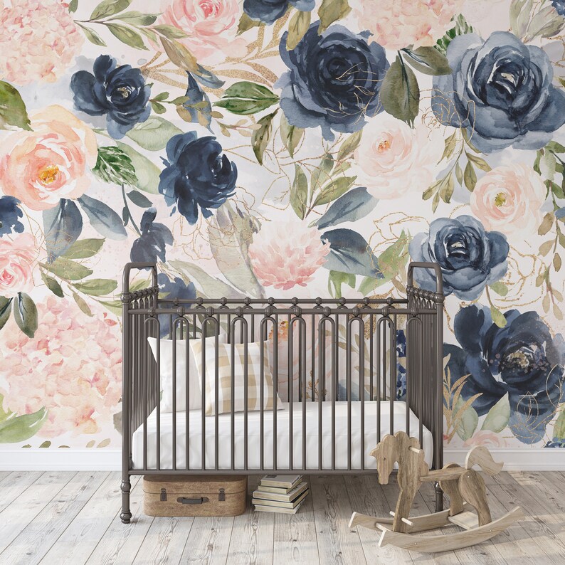 May include: A dark metal crib with vertical bars, a white mattress, and two pillows. A wooden rocking horse and a small suitcase sit on the floor. The wall features a large-scale watercolor floral wallpaper with navy blue and blush pink roses.